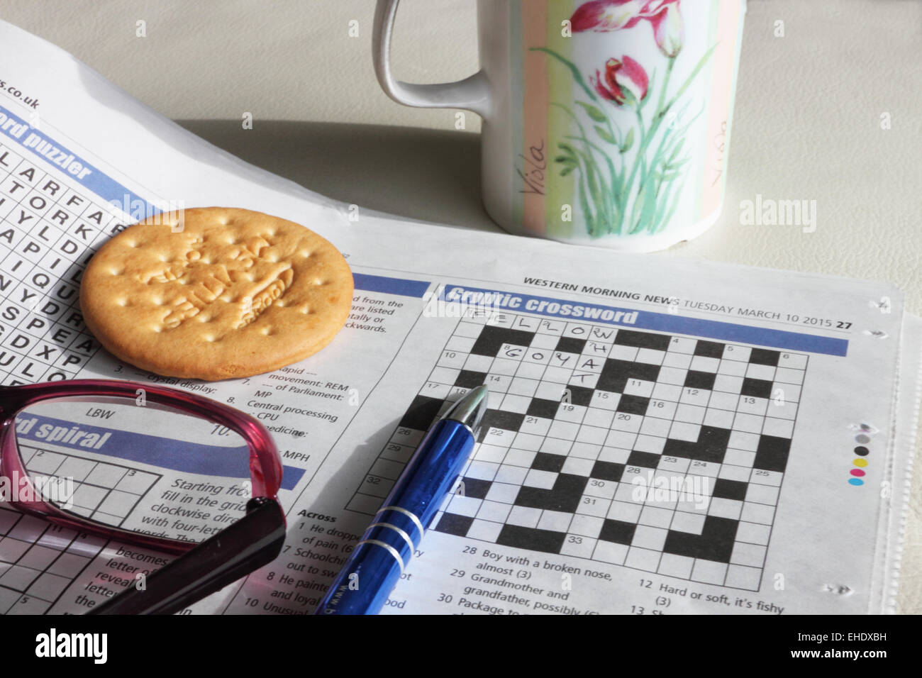 A newspaper open at the crossword page with reading glasses, a pen, mug and biscuit. Stock Photo