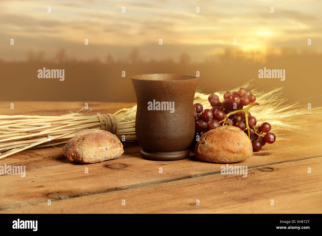 Cup of wine, bread. grapes and wheat on vintage table with warm sunset in background Stock Photo