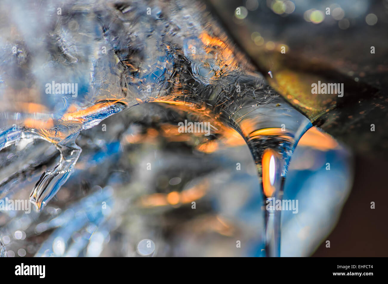 Drop of melting ice water at the drainpipe, with reflections of blue sky and yellow sun rays passing through. Stock Photo