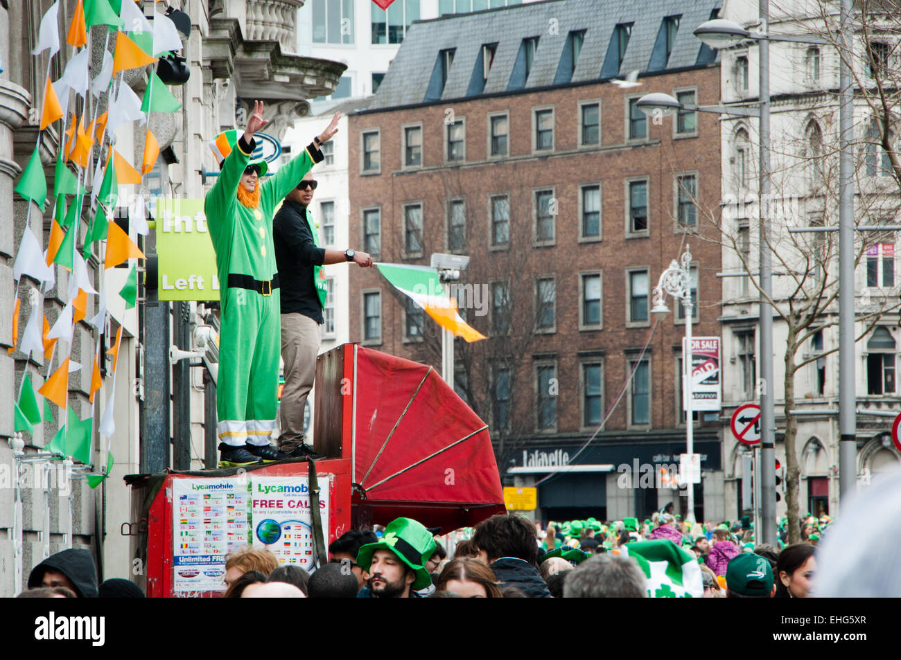 Two men on a news stand above crowds in Dublin city centre on St Patrick's Day wearing a leprechaun suit and with Irish flag Stock Photo