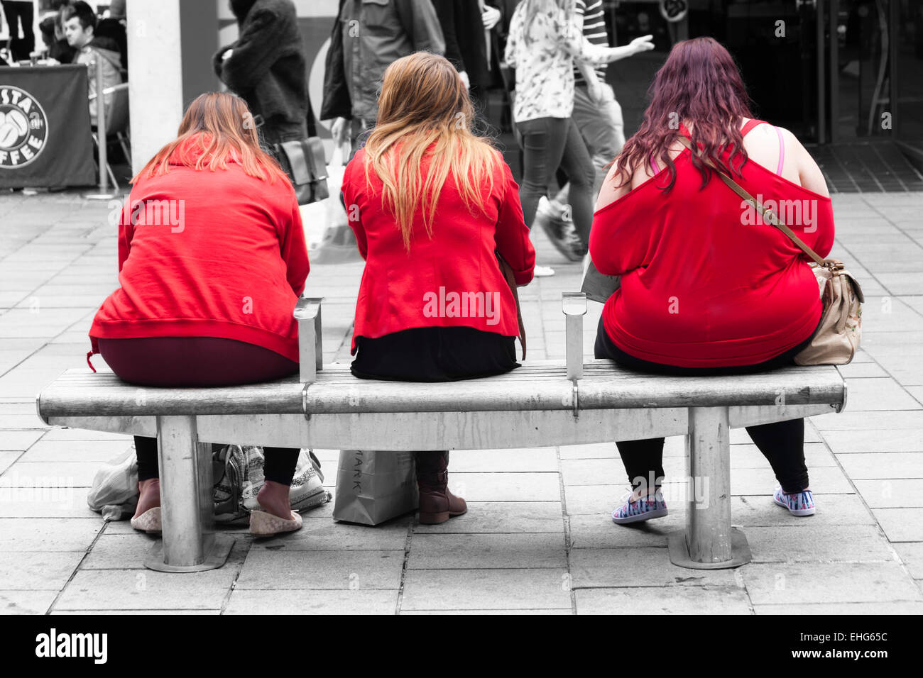 Three young women of different sizes wearing red tops sitting on bench with pedestrians walking past in monochrome at Bristol Stock Photo