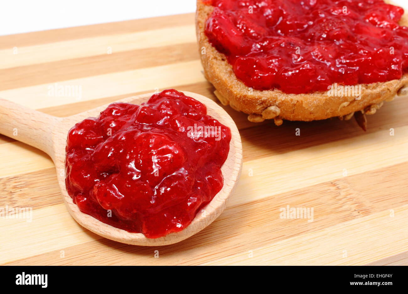 Homemade strawberry jam on wooden spoon and portion of whole wheat bread lying on wooden cutting board, healthy breakfast Stock Photo