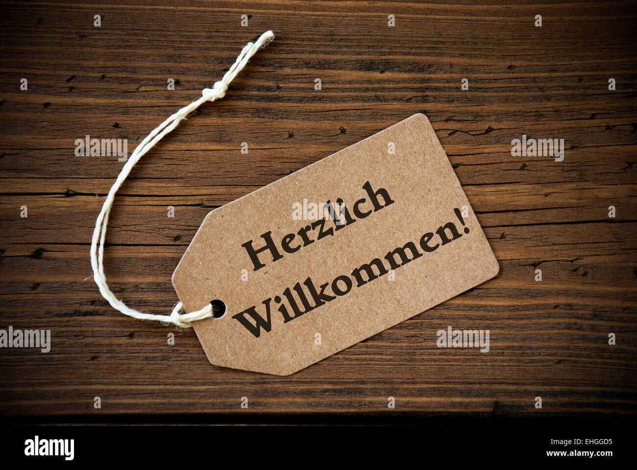 Close Up Of A Brown Label With White Ribbon On Wooden Background With German Text Herzlich Willkommen Frame And Vintage Or Retro Stock Photo