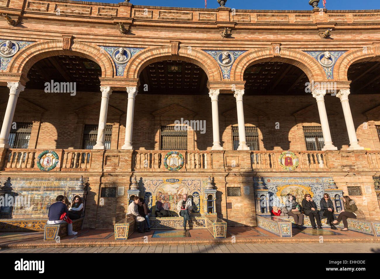 People seating in tiled 'Province Alcoves' along the walls of the Plaza de España, Spain Square, Luisa, in Seville. Stock Photo