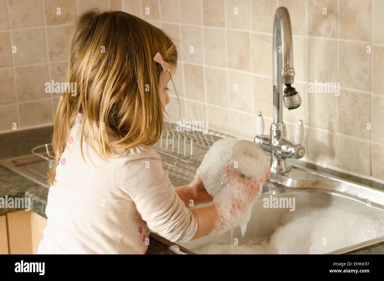 Two-year-old girl playing at washing up at kitchen sink. UK. Stock Photo