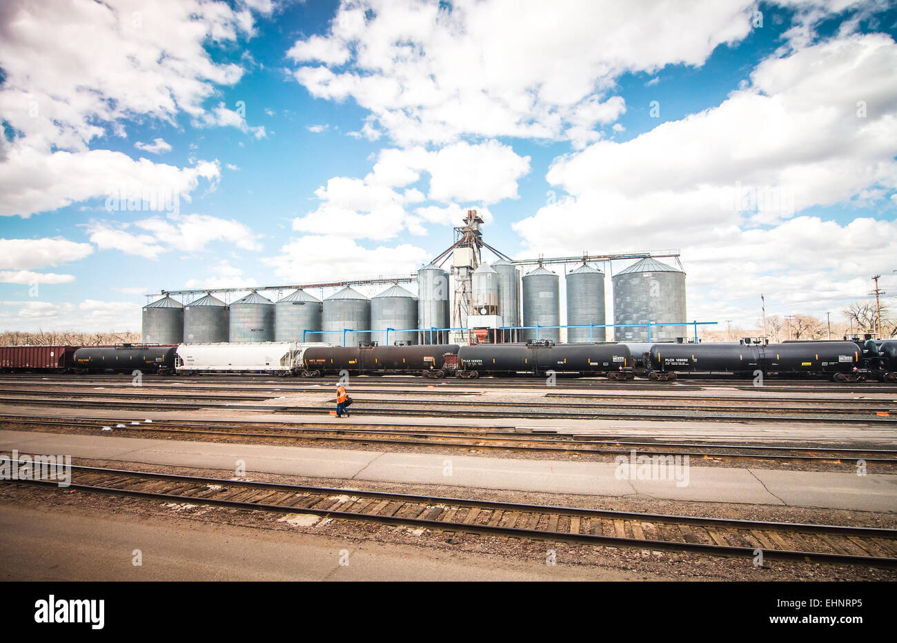 With bright blue skies & open fields beyond, grain silos with rail lines in the foreground represent agriculture in the USA. Stock Photo