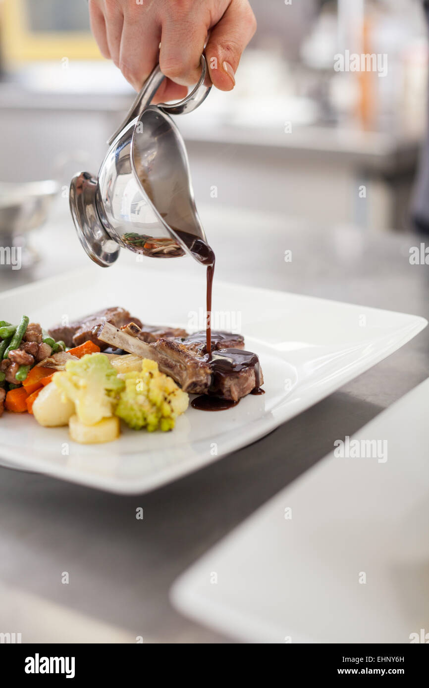 Chef plating up food in a restaurant pouring a gravy or sauce over the meat before serving it to the customer, close up view of Stock Photo