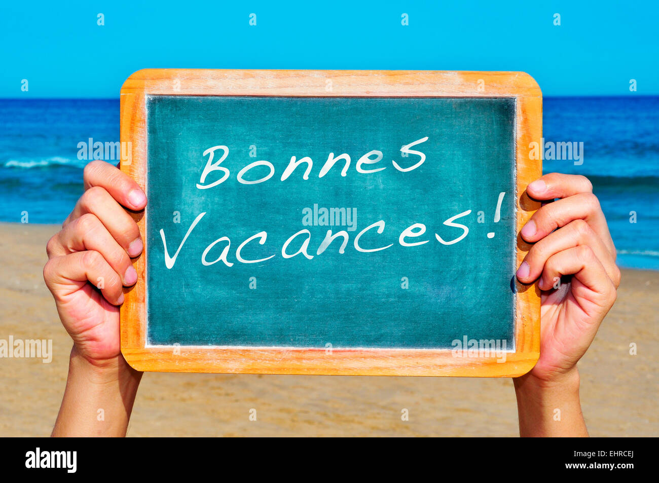 young man holding a blackboard on the beach with the sentenece bonnes vacances, happy vacations in french, written on it Stock Photo