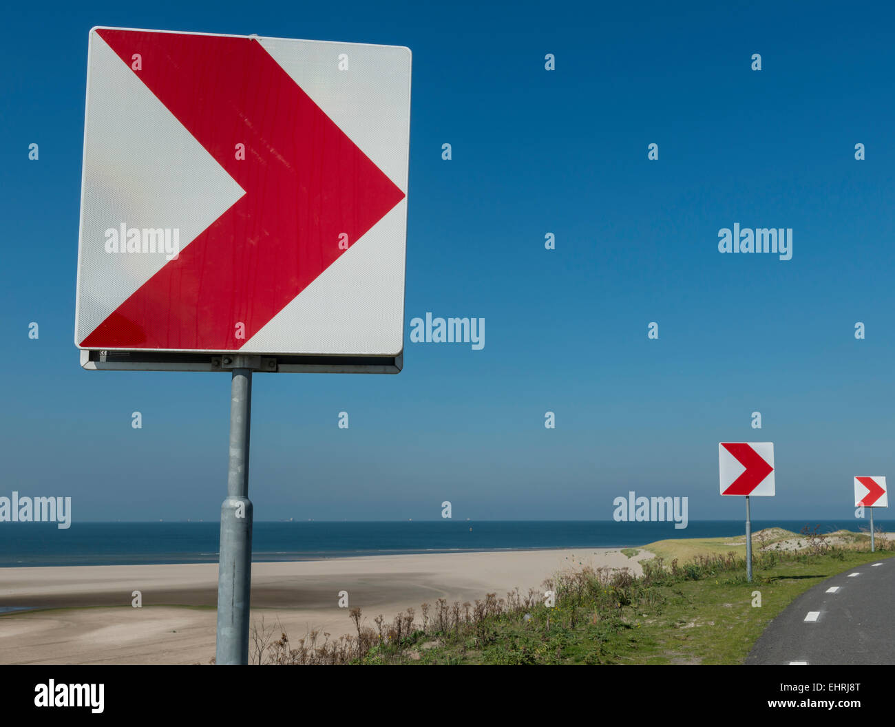 Red and white sign at the road warning for a sharp turn to the right at the coast. Stock Photo