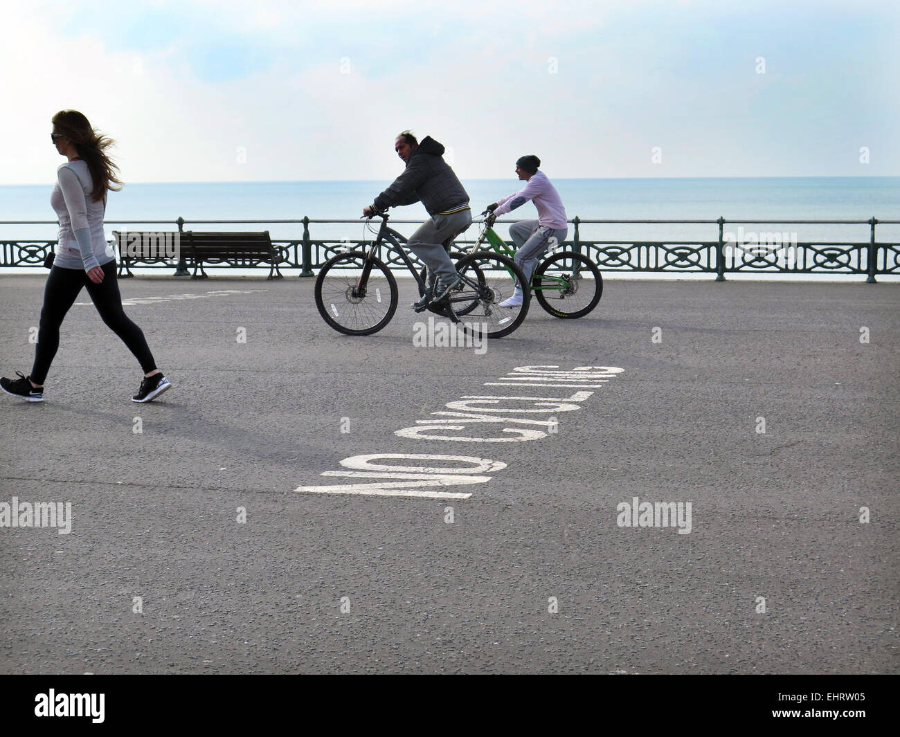 Two male Cyclists, not wearing crash helmets, ignore a NO CYCLING sign painted on the tarmac while cycling and looking at a pretty girl on Hove promenade in the City of Brighton and Hove, East Sussex Stock Photo