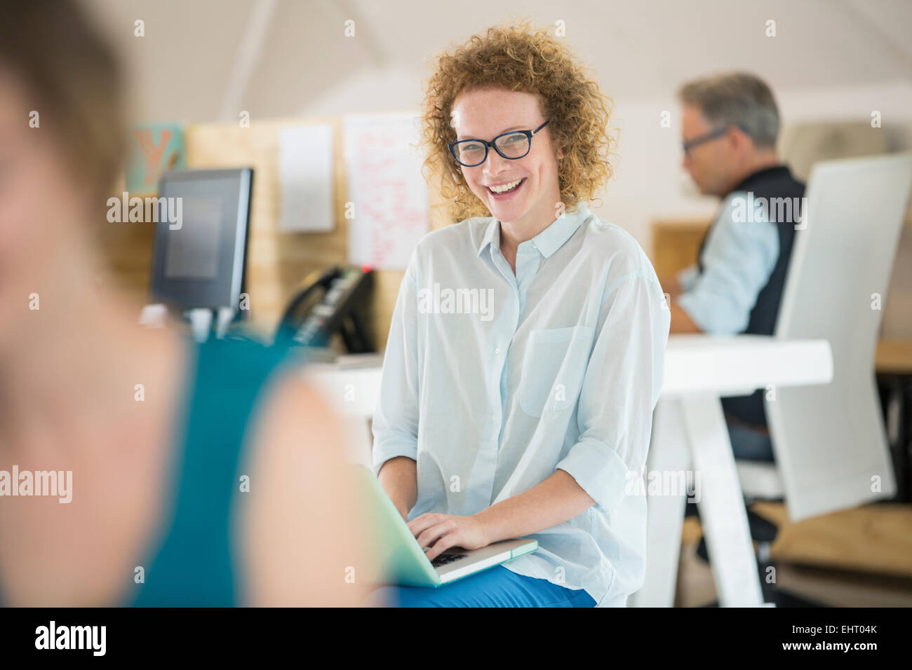 Portrait of woman using laptop and laughing, man working in background Stock Photo