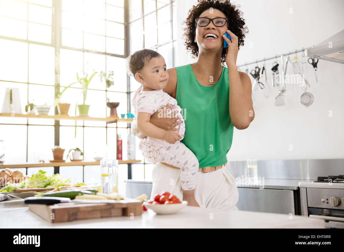 Mother holding her baby daughter while using mobile phone in kitchen Stock Photo