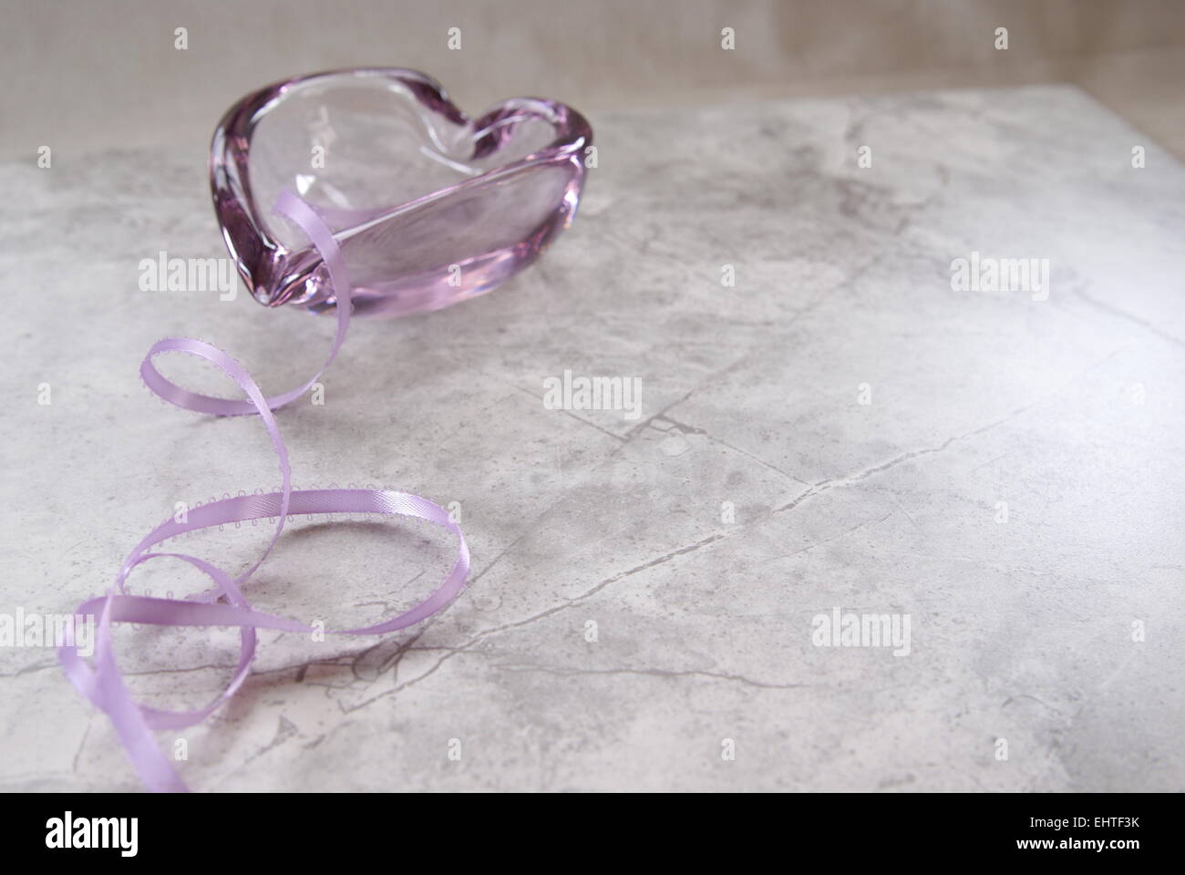 Soft lavender pastel ribbon curls towards a heart shaped light purple, lavender glass dish in the background. Marble surface. Stock Photo