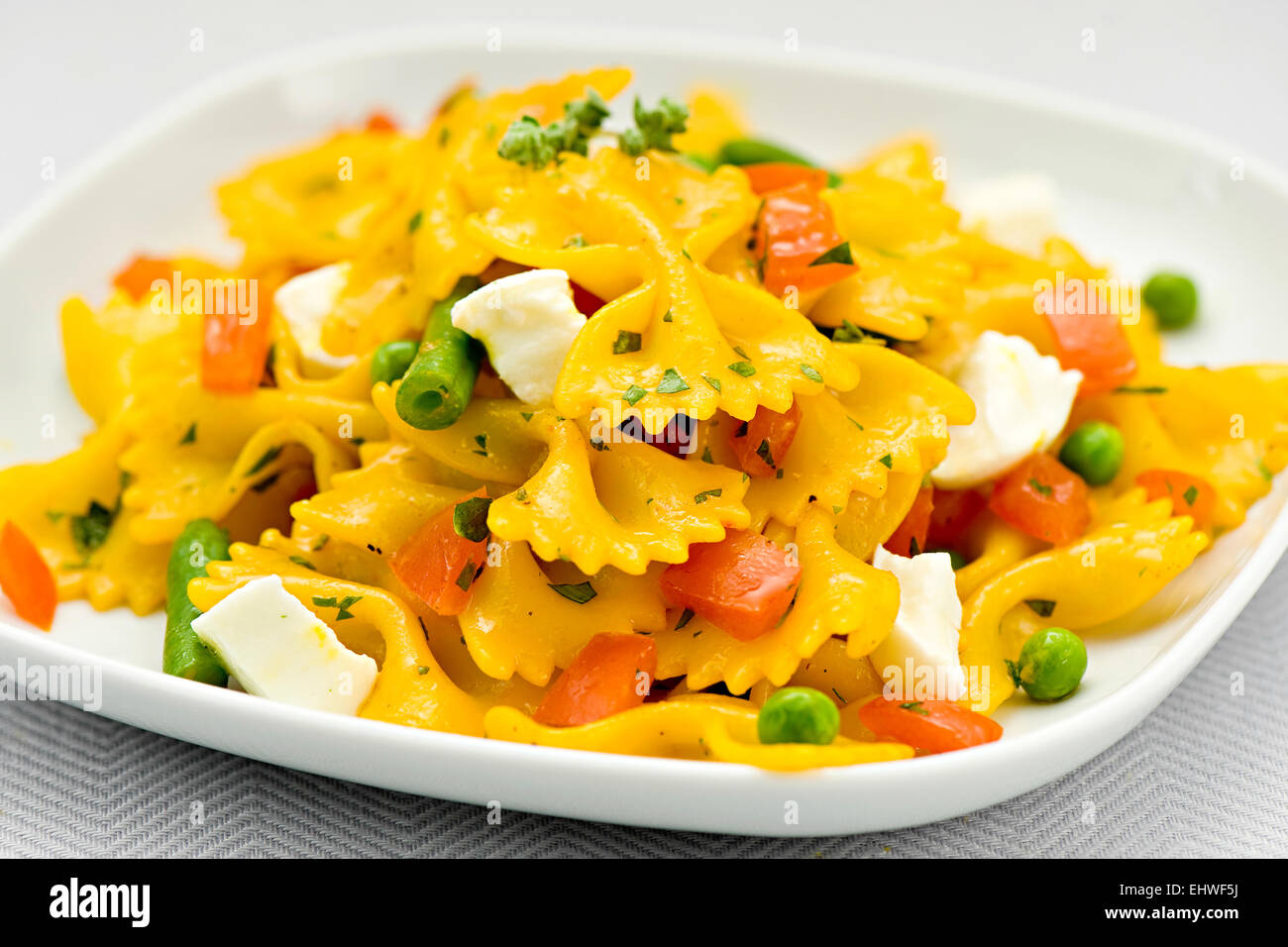 Italian bow tie pasta with mozzarella cheese, fresh tomato, peas and beans sprinkled with chopped herbs served on a square white Stock Photo