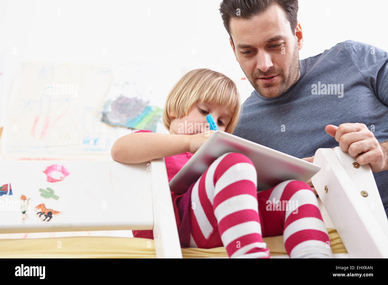 Little girl sitting on bunk bed, drawing on touch pad, father watching Stock Photo