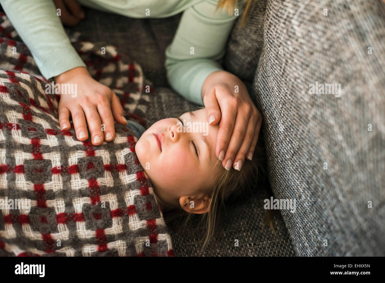 Mother touching forehead of ill daughter lying on couch Stock Photo