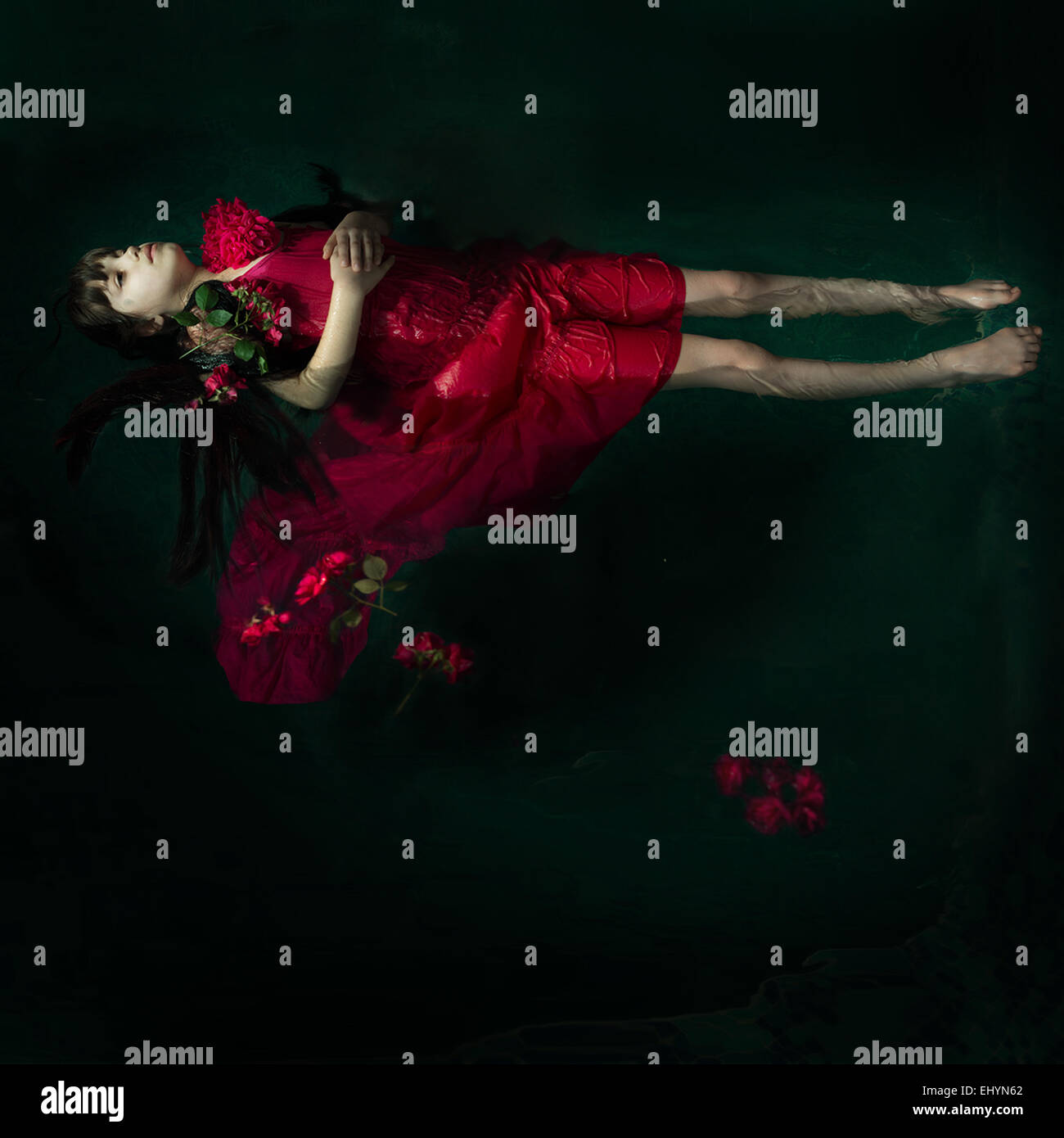 Girl in a red dress floating in water surrounded by red roses Stock Photo