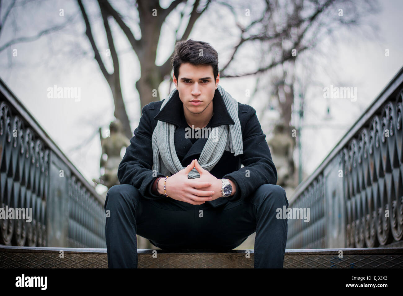 Stylish trendy young man sitting outdoor on old metal bridge or walkway Stock Photo