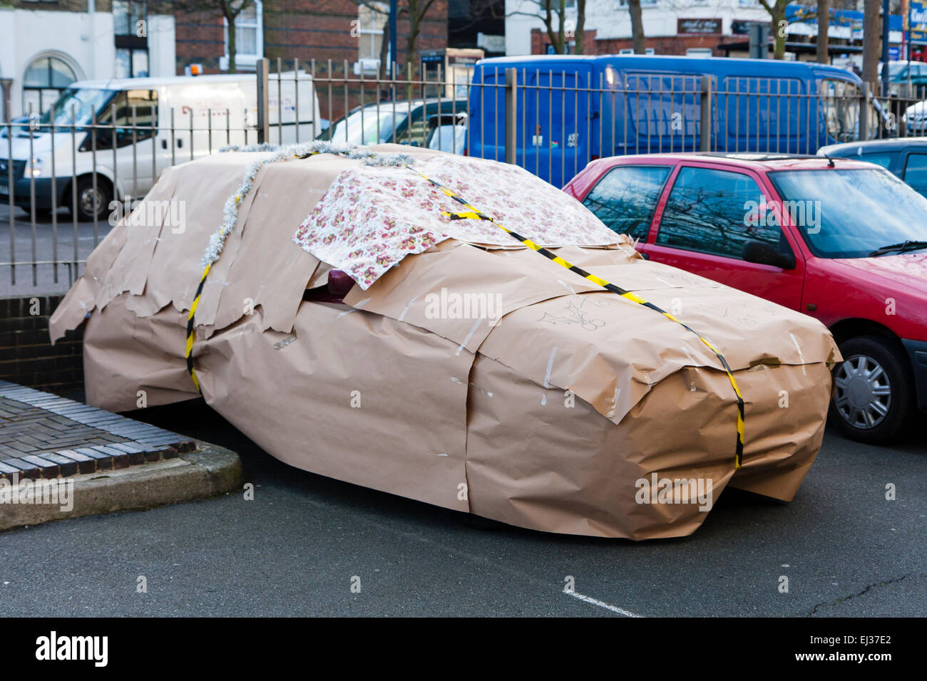 A car wrapped up in brown wrapping paper and black and yellow tape as a surprise secret Santa gift, parked in a car park. Stock Photo