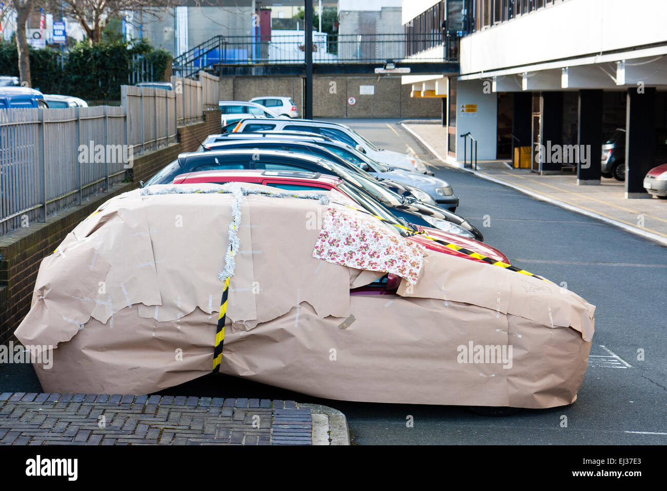 A car wrapped up in brown wrapping paper and black and yellow tape as a surprise secret Santa gift, parked in a car park. Stock Photo