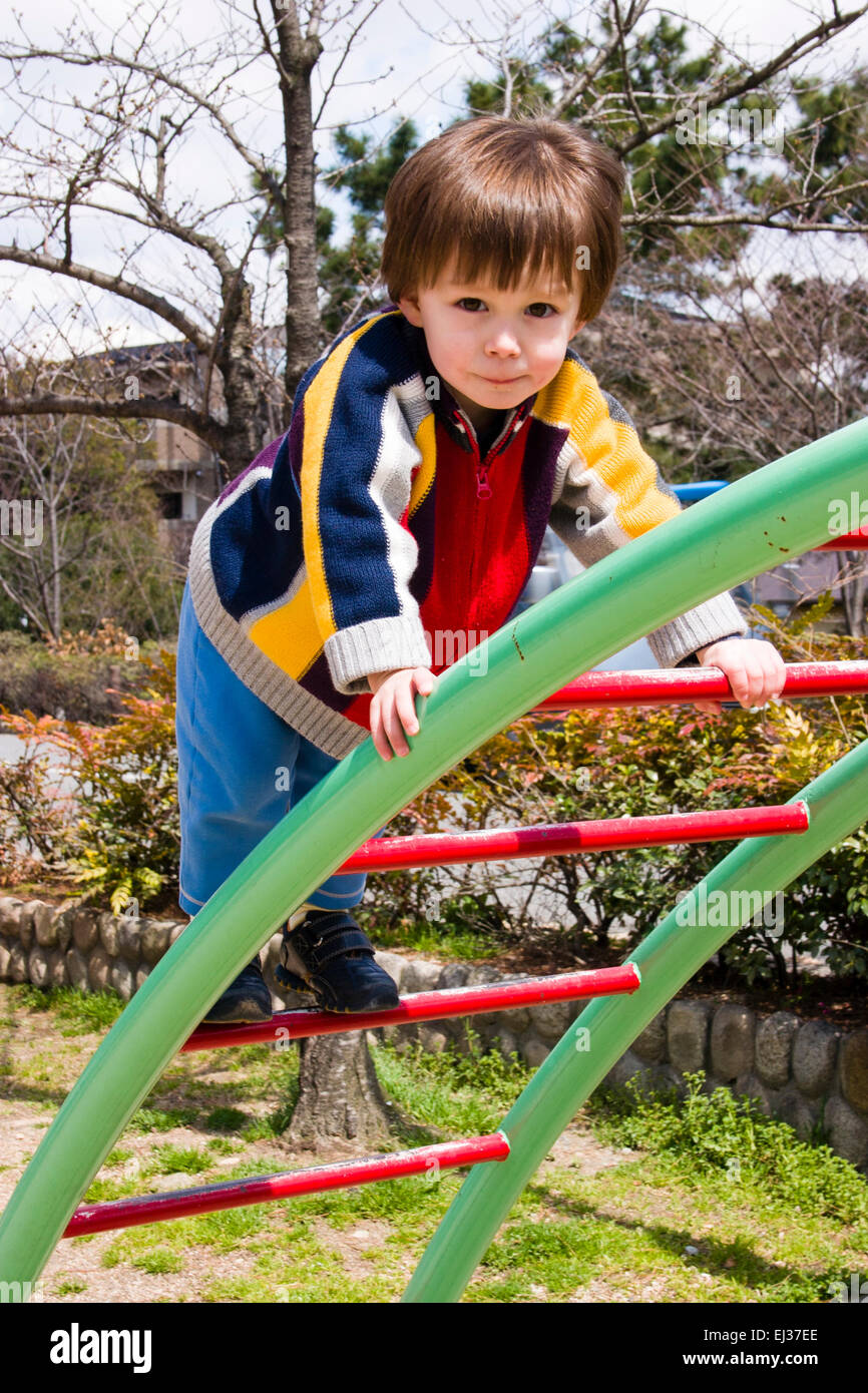 Angelic looking mixed race child, boy, Caucasian Asian, 3-4 year old, standing on climbing frame outdoors in a park, looking very nervous and anxious. Stock Photo