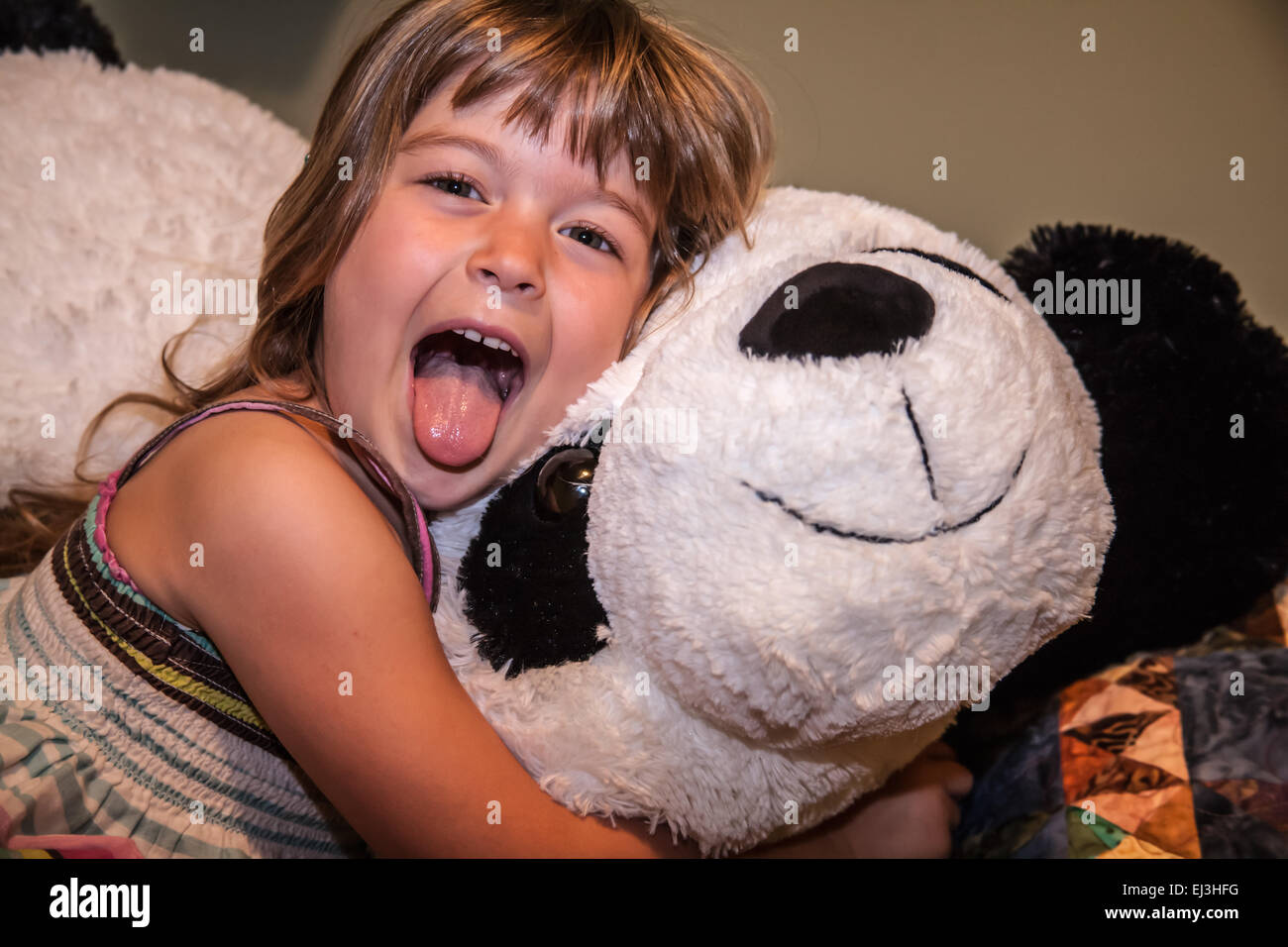 Four year old girl acting up by sticking her tongue out, hugging her large, stuffed panda bear Stock Photo
