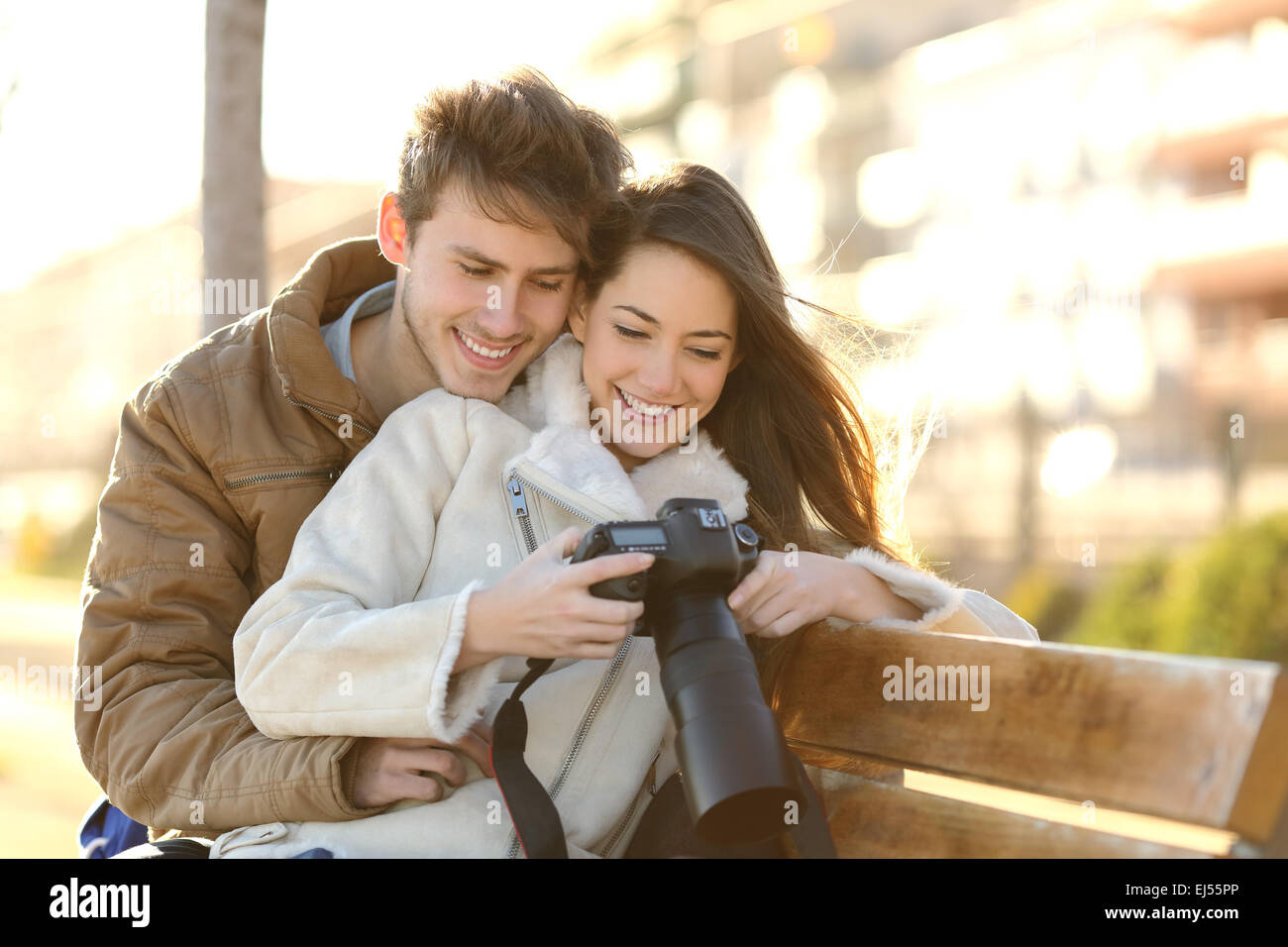 Couple of tourists reviewing photos in a dslr camera sitting in a bench of a park with an urban background Stock Photo