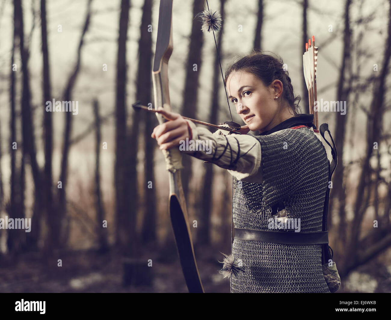 Medieval archer woman, she wearing a chainmail and use a bow and arrow, gloomy forest, cross-processed image. Stock Photo