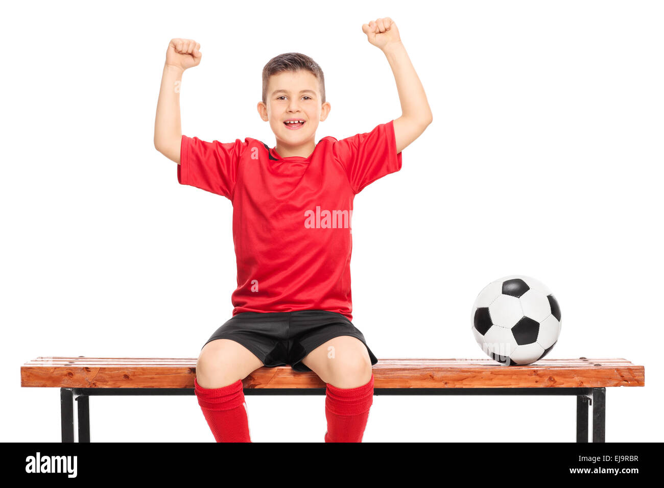 Joyful junior soccer player in red shirt gesturing happiness seated on wooden bench isolated on white background Stock Photo
