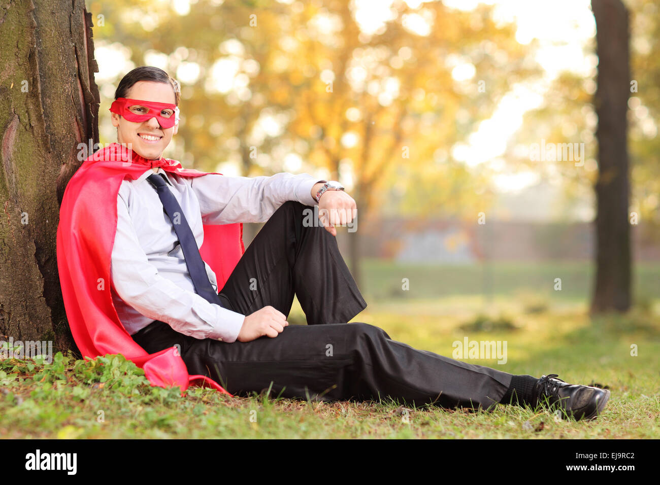 Joyful man in superhero outfit sitting on a grass, leaned on a tree. The shot is in a park during early autumn Stock Photo
