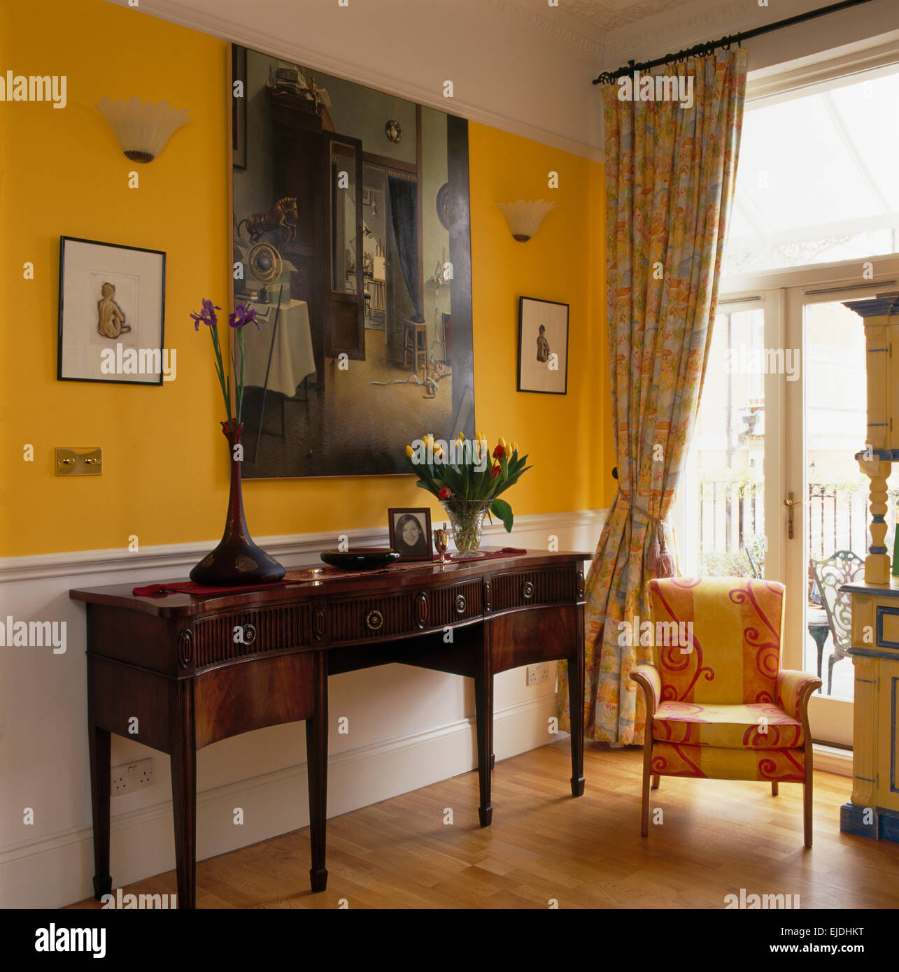 Large painting above antique console table in bright yellow hall with yellow+pink upholstered chair and wooden flooring Stock Photo