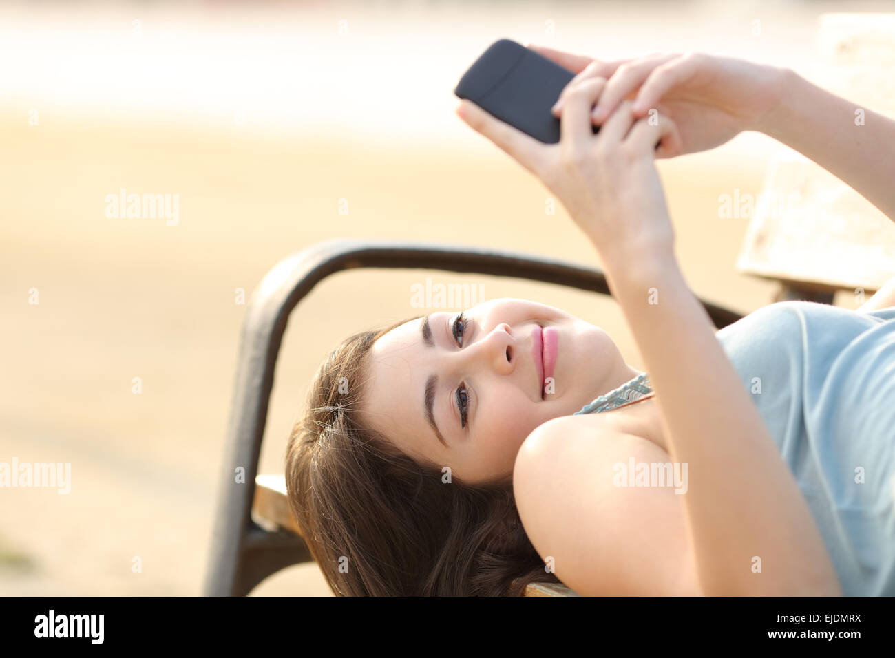 Happy candid teen girl using a smart phone lying in a bench Stock Photo