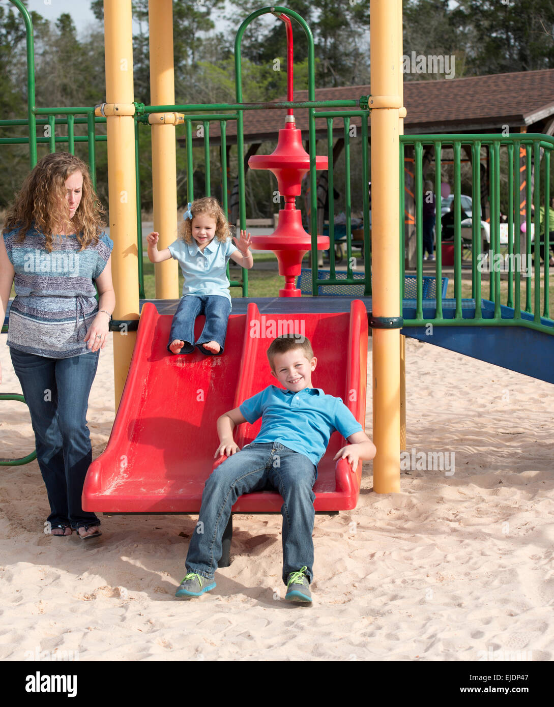 Young boy and girl go down slide on park playground while Mom looks on Stock Photo