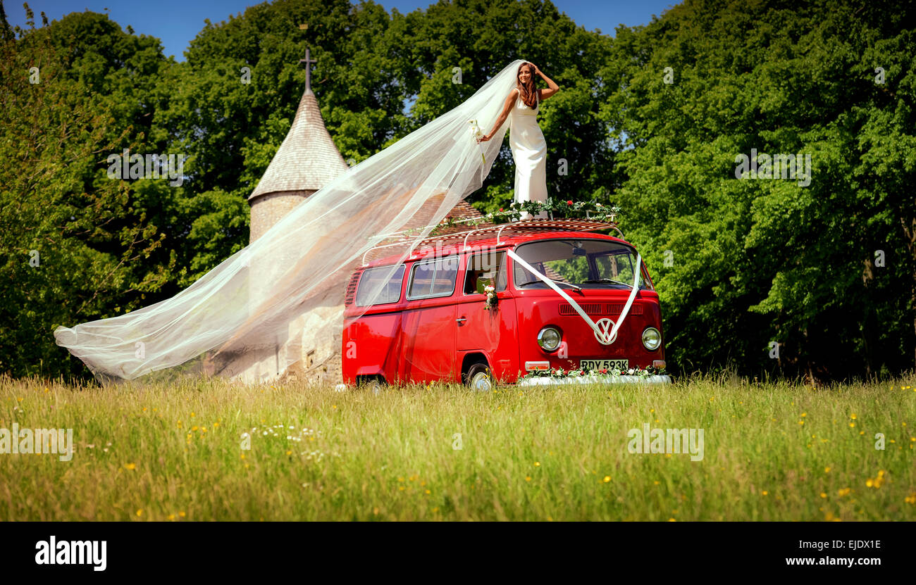 A bride poses on the roof of a bright red VW camper with Church behind. Stock Photo