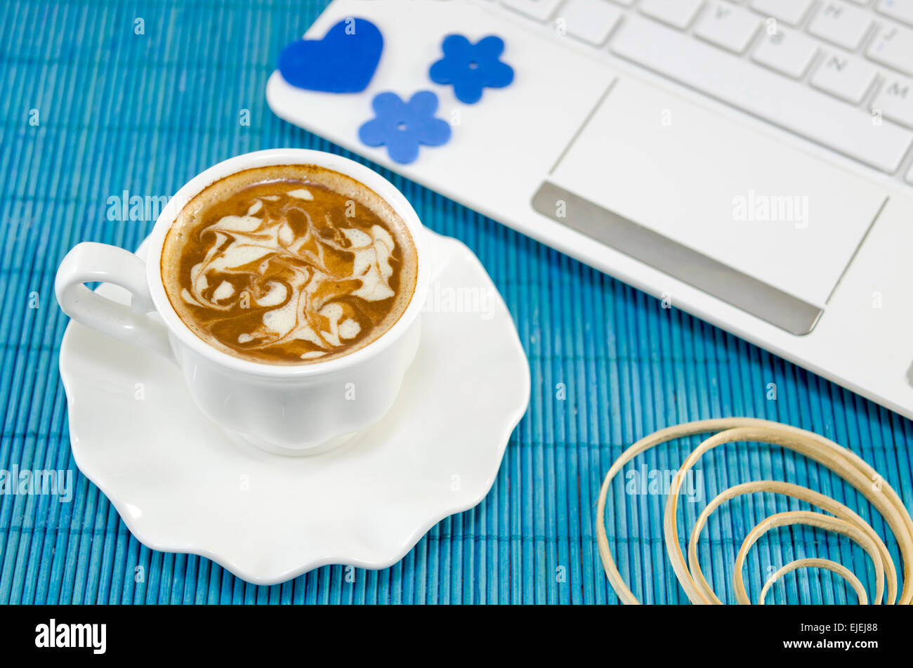 White cup of coffee with decorated foam and a white lap top on a blue tablecloth. Stock Photo