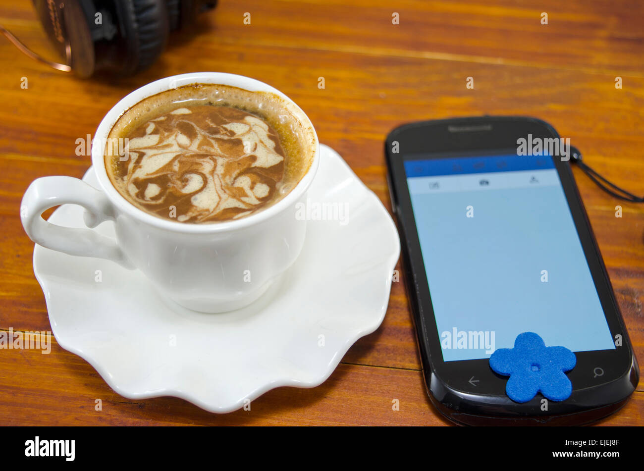 Cup of coffee with decorated foam and a smart phone on a wooden table with a headset in the background Stock Photo