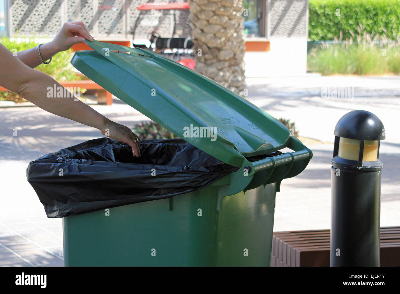 A woman throwing away trash in a large green rubbish bin in a park or