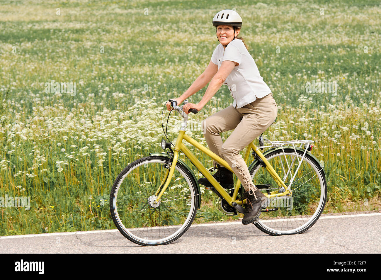 Senior woman vigorously exercising. She's cycling on a country road by the fields. Stock Photo