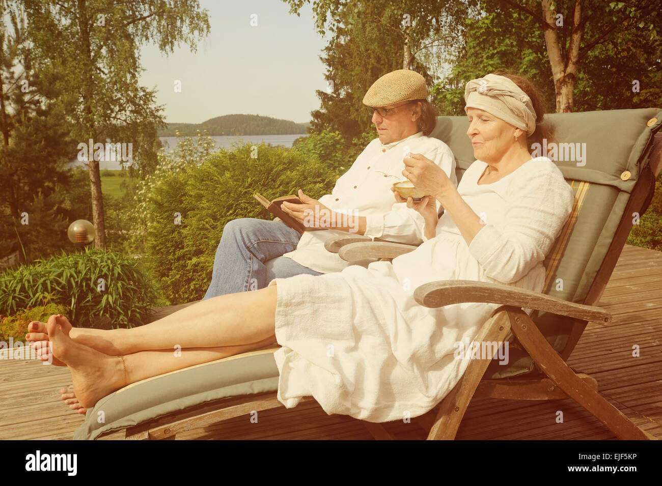 Senior couple relax in deckchairs on a wooden terrace, She is eating from a bowl and he is reading a book. Digital filters used Stock Photo