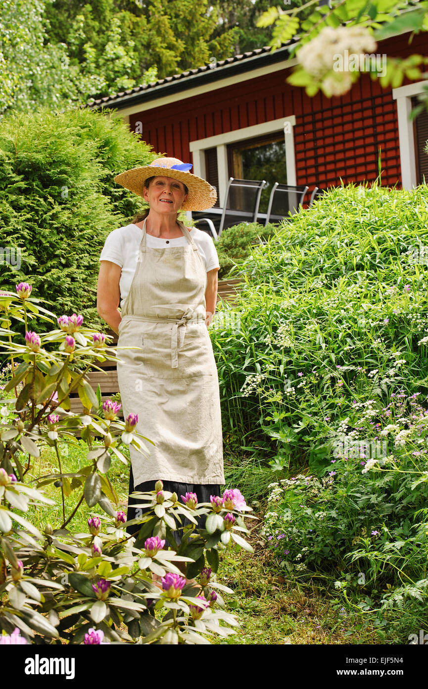 Senior woman standing in her garden, She is surrounded by bushes. There's a red house partly seen in the background. Stock Photo