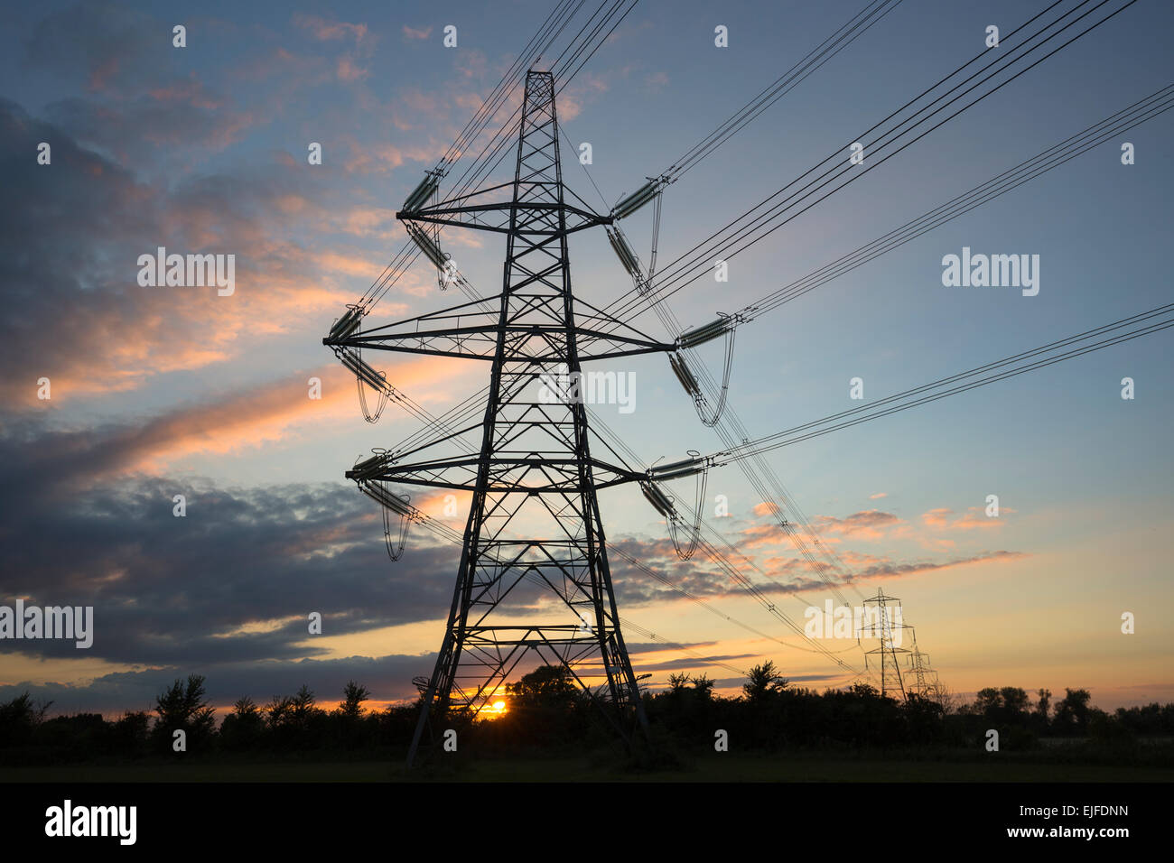 Two forms of energy - setting sun behind electricity pylon and power cables in landscape at sunset in England, UK Stock Photo