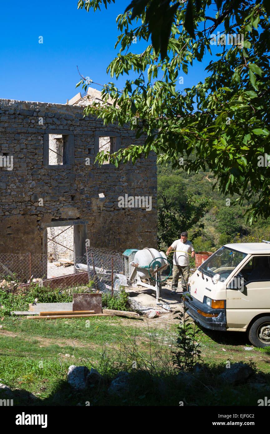 Builder working at cement mixer for masonry restoring derelict house in ruins in re-development in village, Old Perithia - Palea Stock Photo