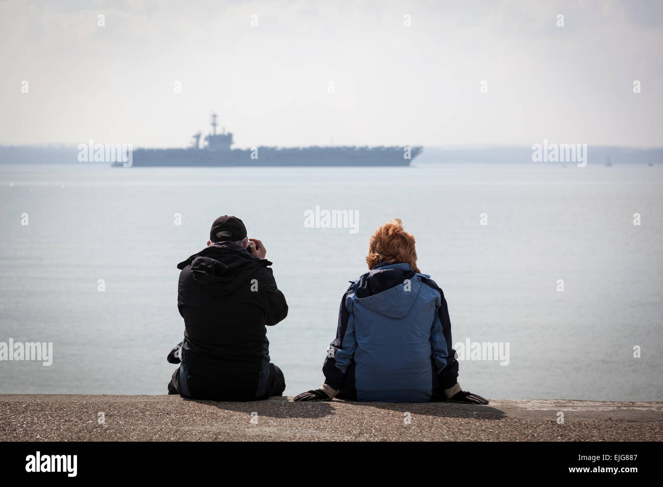 Stokes Bay, Solent, UK. 25th March, 2015. A couple sit on a concrete path at Stokes Bay whilst the man photographs the Nimitz class aircraft carrier USS Theodore Roosevelt at anchor in the Solent, 25th March 2015. Credit:  Anthony Hatley/Alamy Live News Stock Photo