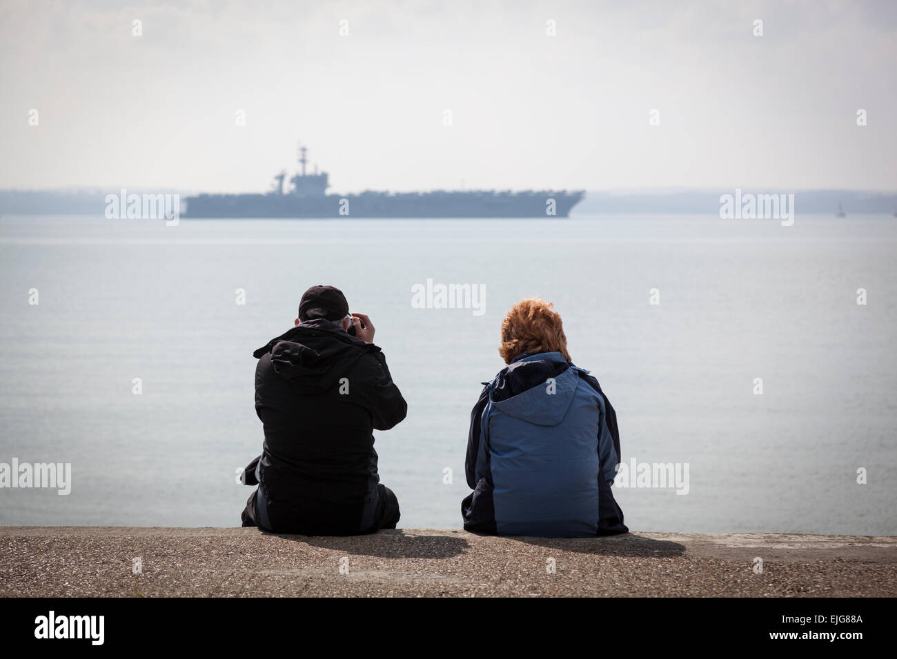 Stokes Bay, Solent, UK. 25th March, 2015. A couple sit on a concrete path at Stokes Bay whilst the man photographs the Nimitz class aircraft carrier USS Theodore Roosevelt at anchor in the Solent, 25th March 2015. Credit:  Anthony Hatley/Alamy Live News Stock Photo