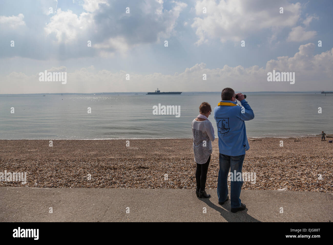 Stokes Bay, Solent, UK. 25th March, 2015. A couple survey the Solent with binoculars as the Nimitz class aircraft carrier USS Theodore Roosevelt lies at anchor in the distance, 25th March 2015. Credit:  Anthony Hatley/Alamy Live News Stock Photo