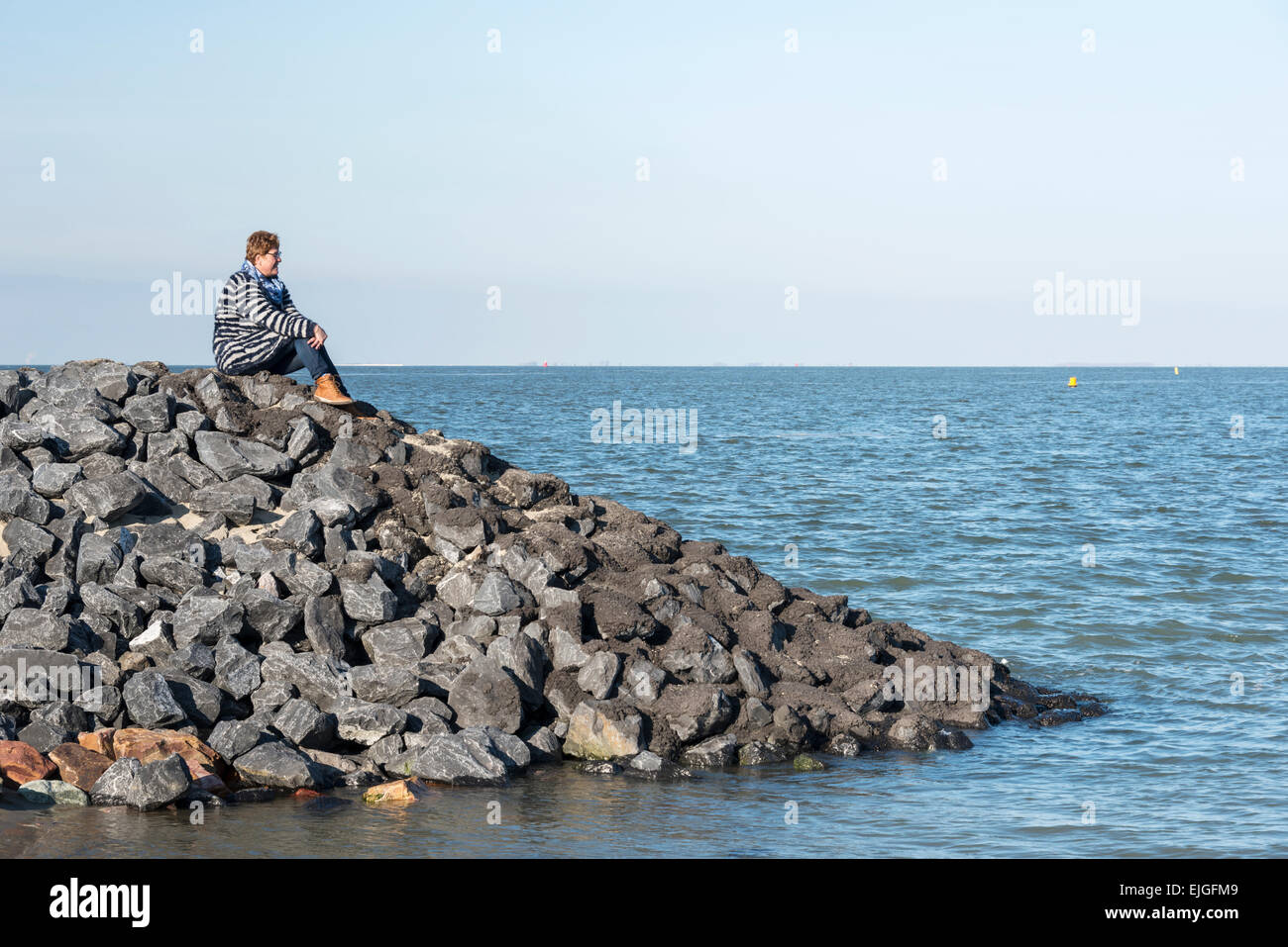 adult white woman with brown boots and blue clothes sitting on the rocks near the sea and looking to the horizon waiting for som Stock Photo
