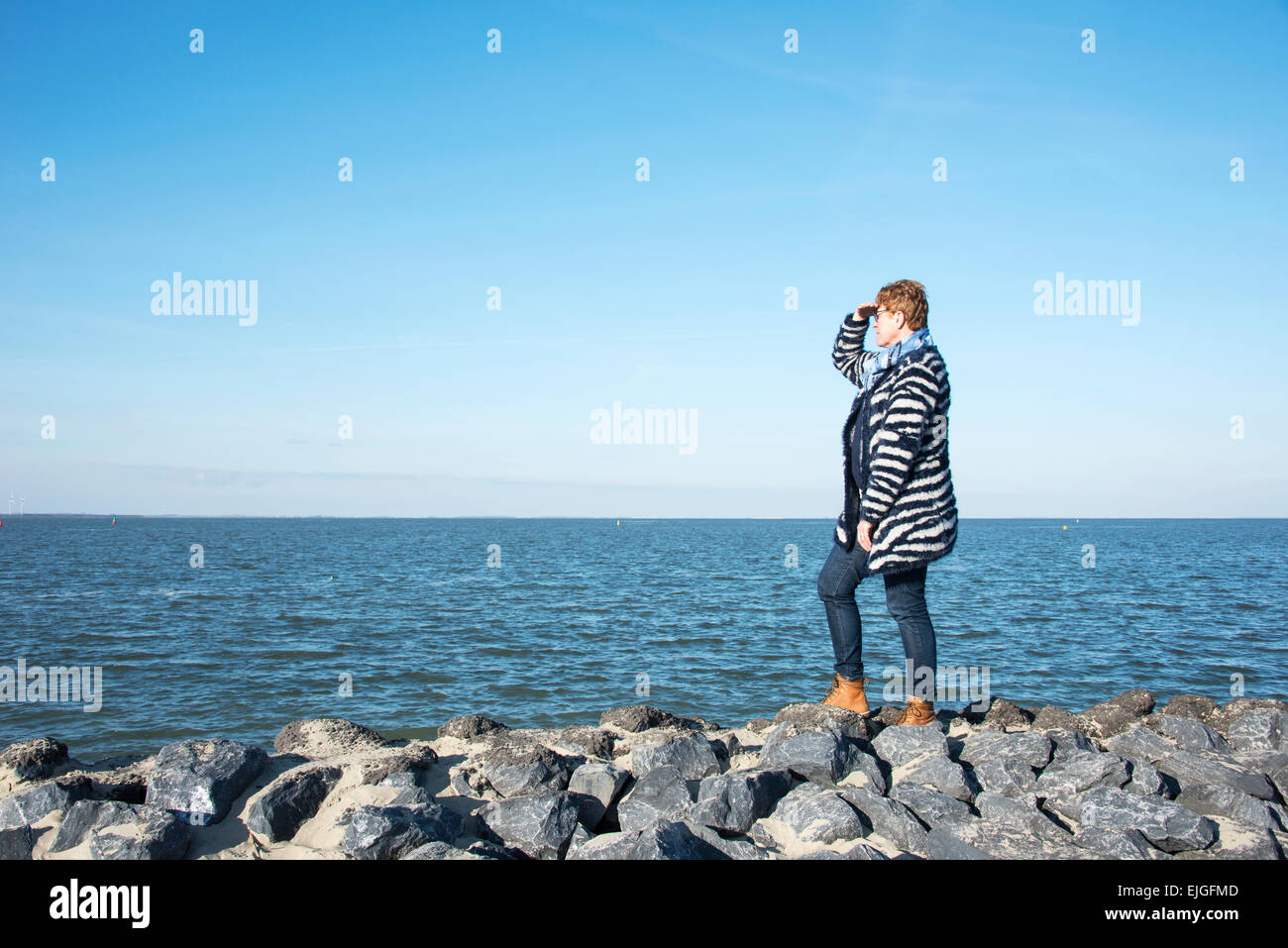 adult woman in blue clothes and brown boots standing on the rocks at the coast and staring to the horizon over the empty sea Stock Photo