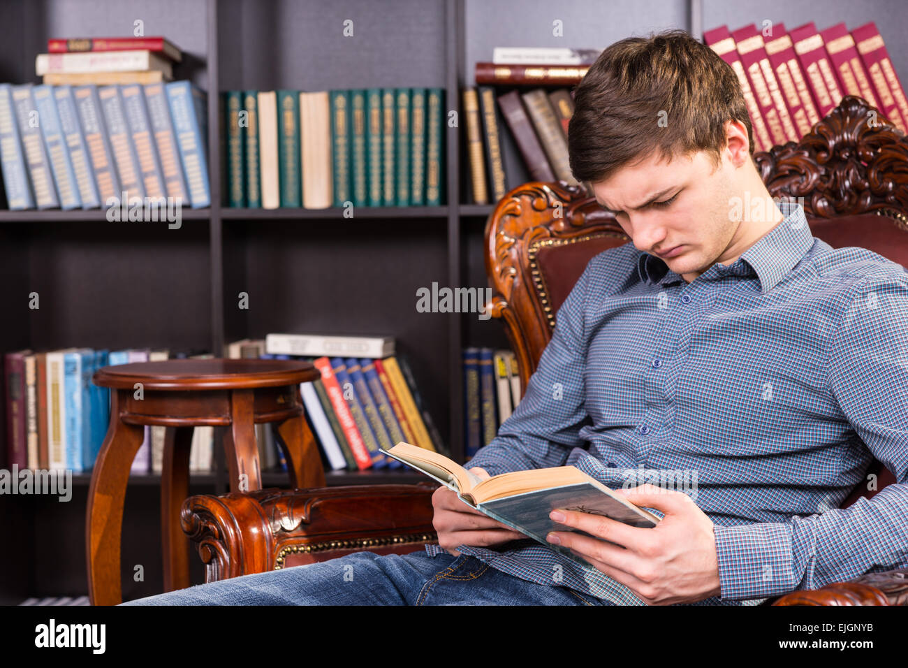 Handsome young man sitting in a comfortable antique carved wooden armchair reading a book in a library Stock Photo