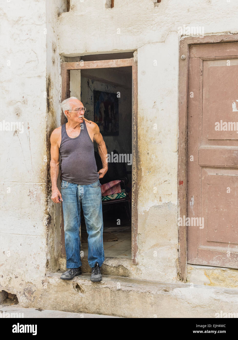 Hispanic Cuban senior citizen man with gray hair stands in the doorway of his home smoking a cigar. Stock Photo