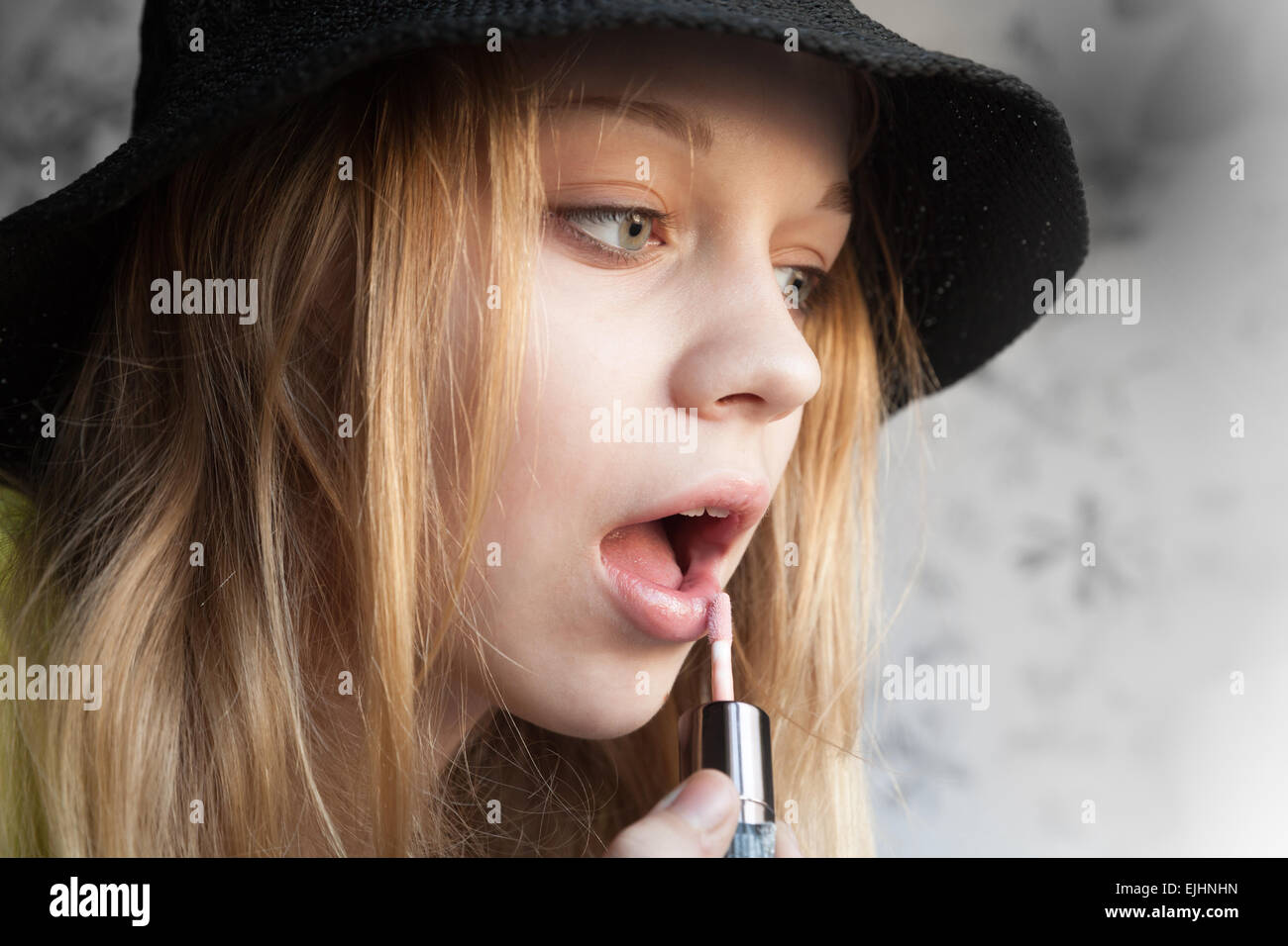 Portrait of beautiful blond teenage girl in black hat doing make up with liquid lipstick Stock Photo
